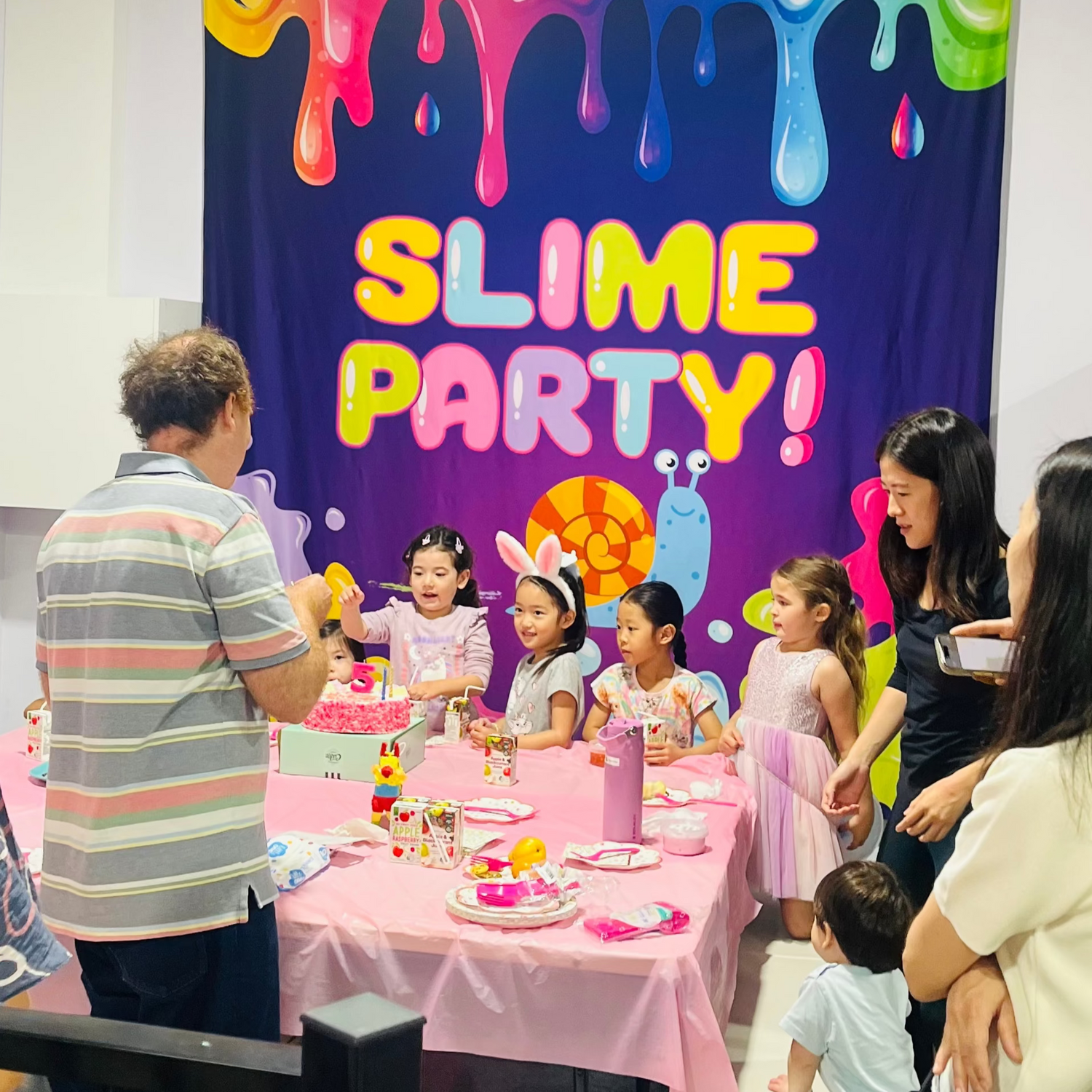 Children's slime party with children and adults gathered around a table, decorated with a 'Slime Party!' banner at Slime Mania brisbane
