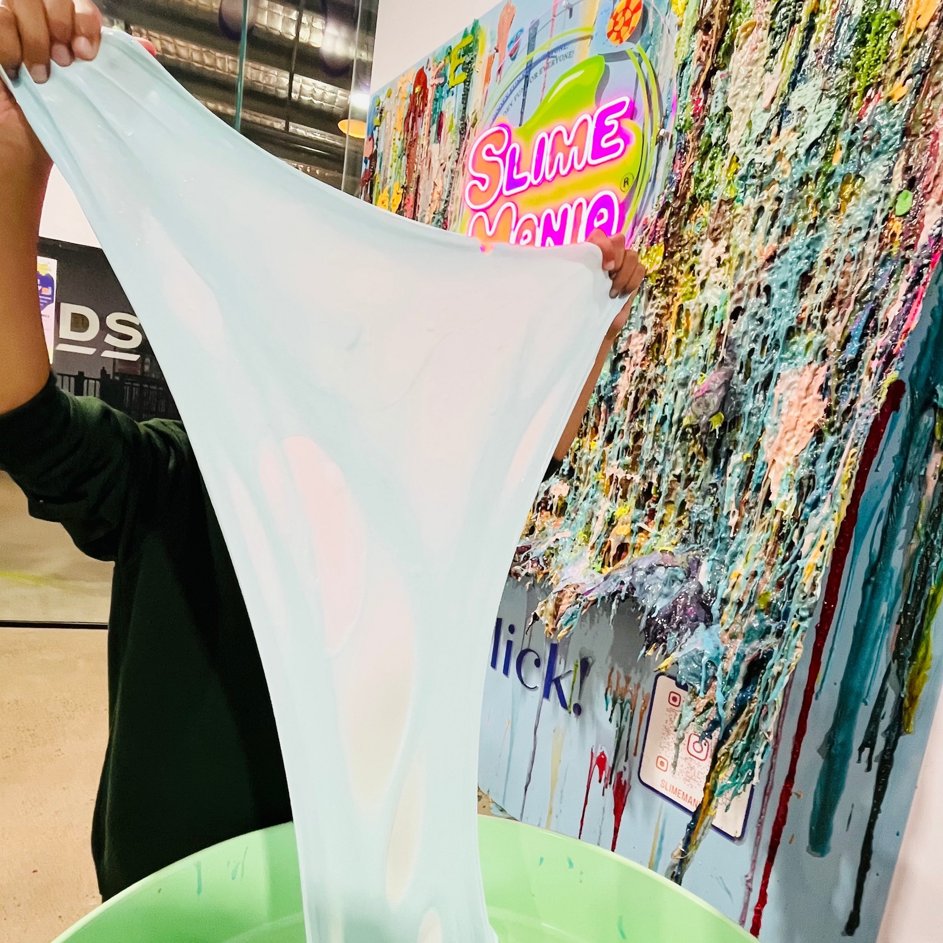 Person stretching a large sheet of baby blue slime over a green tray with a colorful slime wall in the background at Slime Mania brisbane